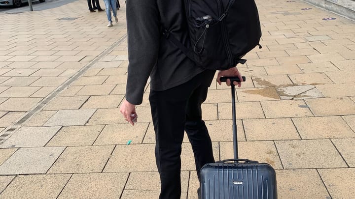 Lee from the shoulders down on Princes Street in Edinburgh, Tom Bihn backpack on, hand on his Rimowa carry-on